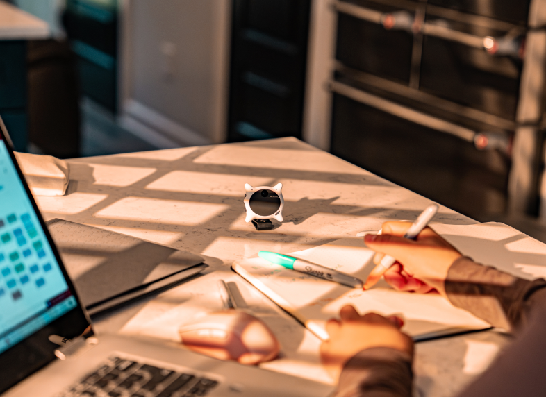 A white Looki L1 stands on a sunlit desk while someone writes notes beside a laptop.